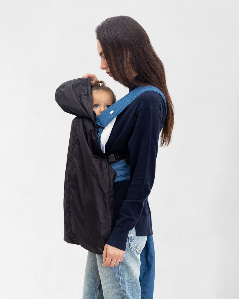 דוגמנית עם תינוק במנשא מכוסה במעיל מנשא לתינוק בצבע שחור עשוי שכבה חיצונית של ניילון ושכבה פנימית מפליז, כיסוי מנשא לתינוק לחורף במקום שמיכת מנשא