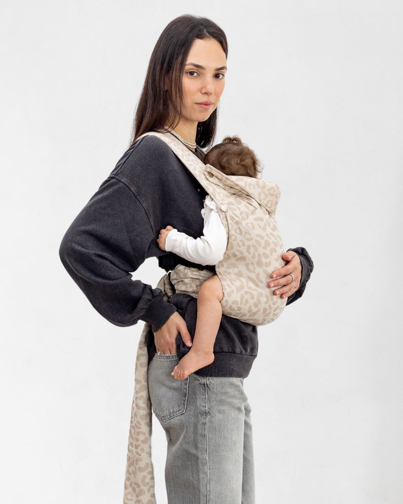 דוגמנית בתמונת פרופיל נושאת תינוקת בת 11 חודשים במנשא ילקוט מגיל לידה בצבע אופוויט מבד פשתן טבעי עם הדפס מנומר של חברת אורבן בייבי ראפ במנח בטן לבטן  בעיצוב דומה לחברת ארטיפופ