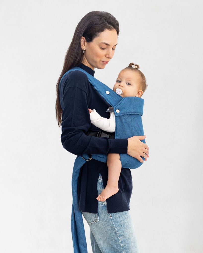 דוגמנית בתמונה צדדית נושאת תינוקת בת 11 חודשים במנשא ילקוט מגיל לידה בצבע ג'ינס של חברת אורבן בייבי ראפ