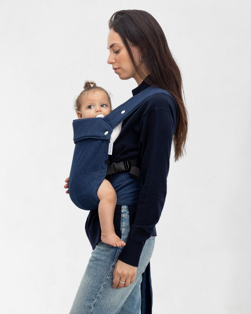 דוגמנית בתמונת פרופיל נושאת תינוקת בת 11 חודשים במנשא ילקוט מגיל לידה בצבע ג'ינס כהה של חברת אורבן בייבי ראפ