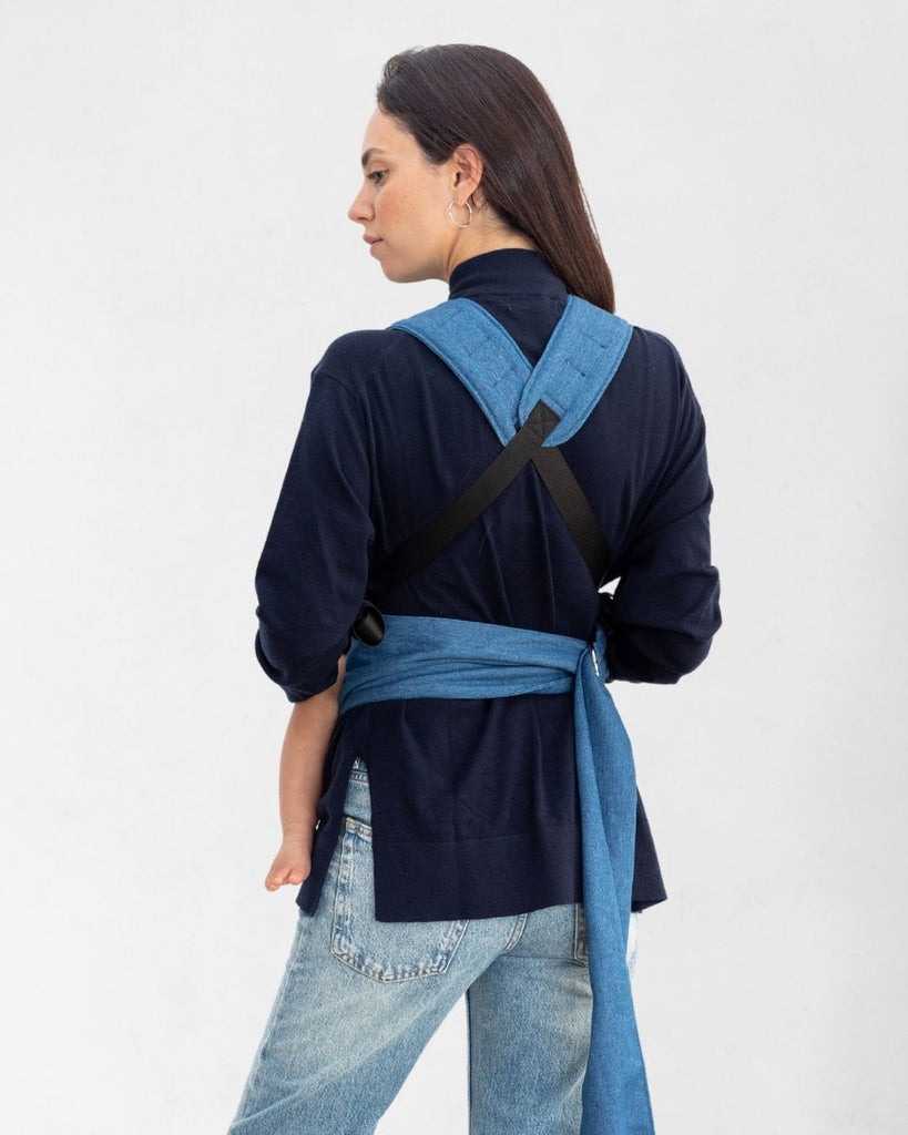 דוגמנית בתמונת גב נושאת תינוקת בת 11 חודשים במנשא ילקוט מגיל לידה בצבע ג'ינס של חברת אורבן בייבי ראפ עם רצועות מוצלבות על הגב 