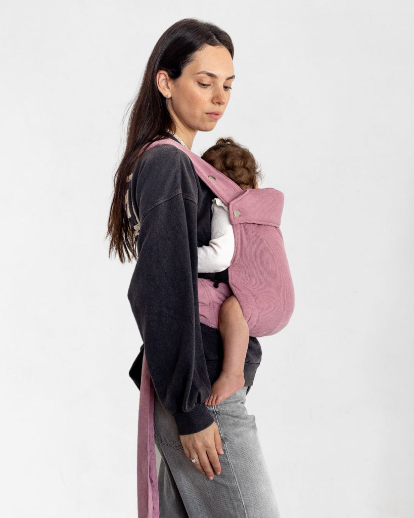 דוגמנית בתמונת פרופיל נושאת תינוקת בת 11 חודשים במנשא ילקוט מגיל לידה בצבע ורוד עתיק מבד קורדרוי דק טבעי של חברת אורבן בייבי ראפ במנח בטן לבטן  בעיצוב דומה לחברת ארטיפופ