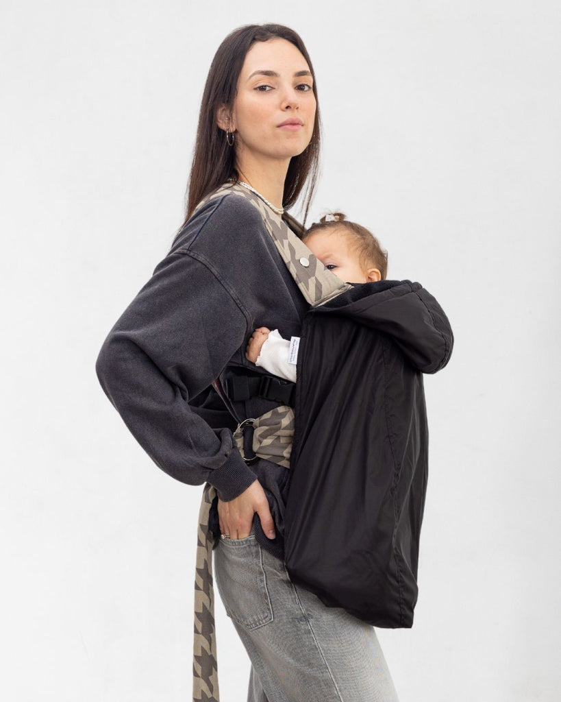 דוגמנית עם תינוק במנשא מכוסה במעיל מנשא לתינוק בצבע שחור עשוי שכבה חיצונית של ניילון ושכבה פנימית מפליז, כיסוי מנשא לתינוק לחורף במקום שמיכת מנשא