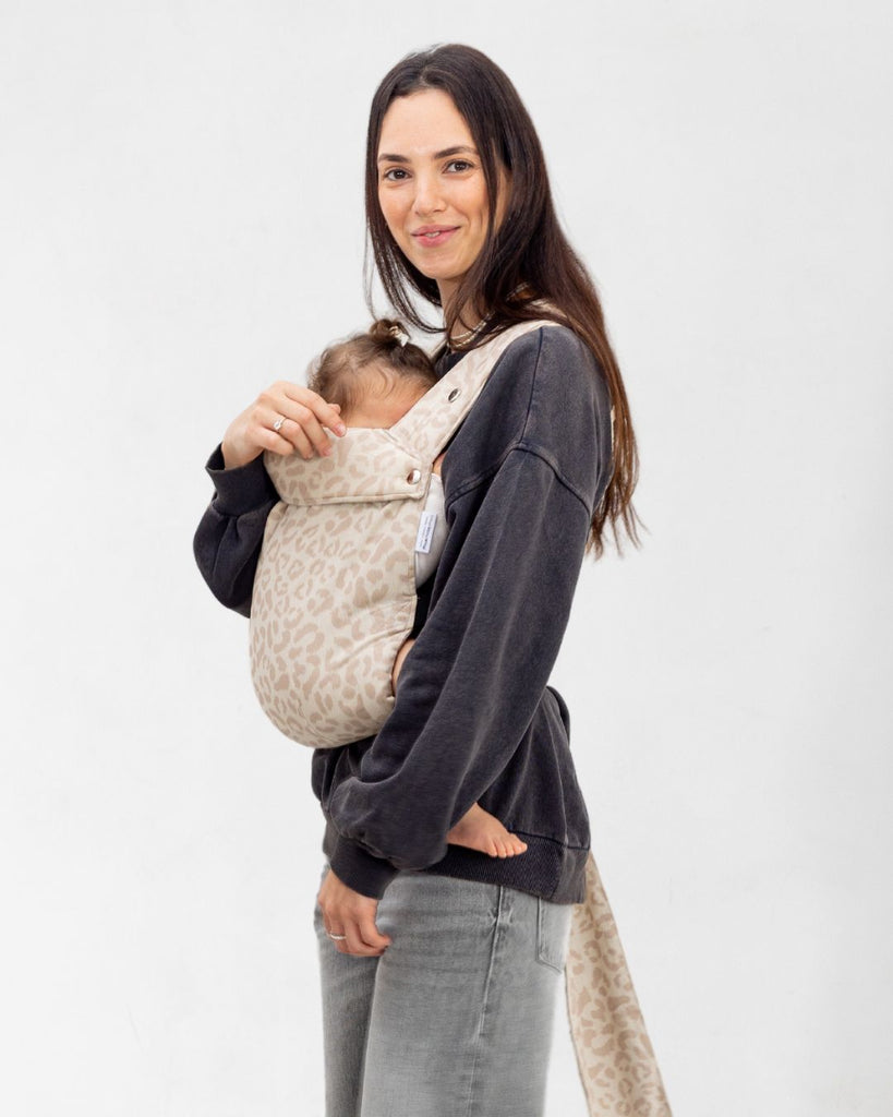 דוגמנית בתמונת פרופיל נושאת תינוקת בת 11 חודשים במנשא ילקוט מגיל לידה בצבע אופוויט מבד פשתן טבעי עם הדפס מנומר של חברת אורבן בייבי ראפ במנח בטן לבטן  בעיצוב דומה לחברת ארטיפופ