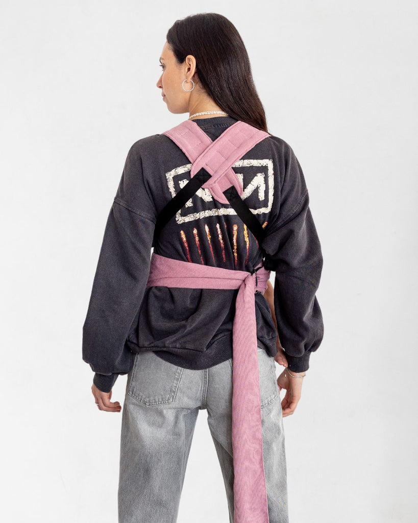 דוגמנית בתמונת גב נושאת תינוקת בת 11 חודשים במנשא ילקוט מגיל לידה בצבע ורוד עתיק מבד קורדרוי דק טבעי של חברת אורבן בייבי ראפ במנח בטן לבטן  בעיצוב דומה לחברת ארטיפופ עם רצועות מוצלבות באיקס על הגב 