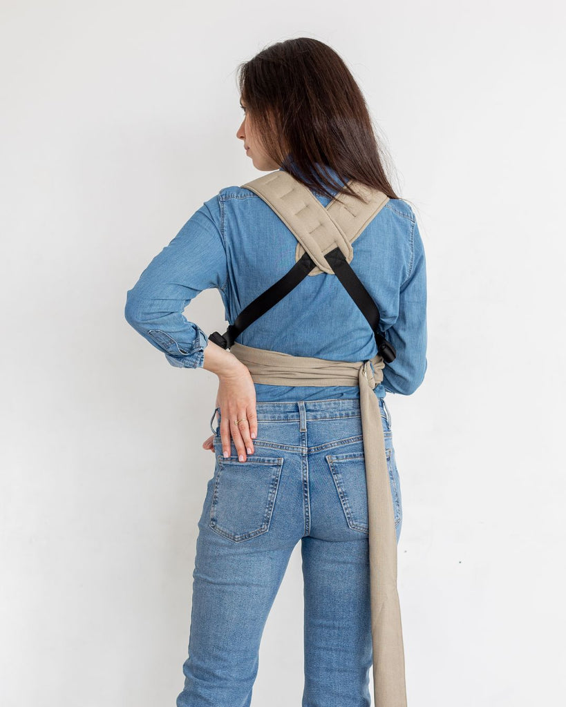 דוגמנית בתמונת גב נושאת תינוקת בת 5 חודשים במנשא ילקוט מגיל לידה בצבע בז' מבד פשתן טבעי של חברת אורבן בייבי ראפ במנח בטן לבטן  עם רצועות מוצלבות באיקס על הגב 