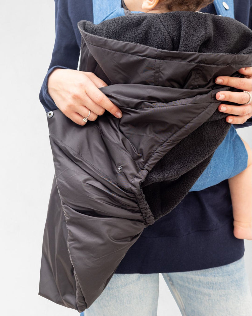 דוגמנית עם תינוק במנשא מכוסה במעיל מנשא לתינוק בצבע שחור עשוי שכבה חיצונית של ניילון ושכבה פנימית מפליז, כיסוי מנשא לתינוק לחורף במקום שמיכת מנשא